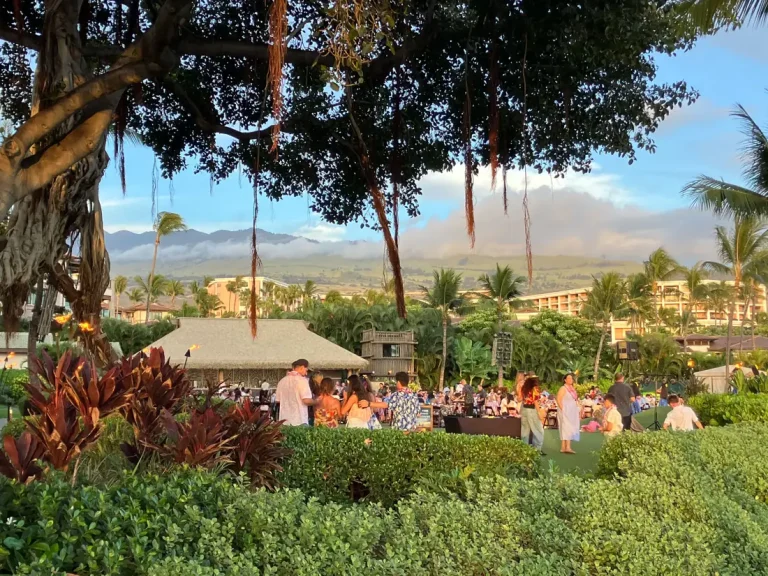 Guests gathering on a resort lawn for a Maui luau at golden hour, with palm trees and mountains behind