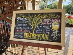 Chalkboard sign at Monkeypod Kitchen listing happy hour from 3:30 to 5PM, live music and dinner times