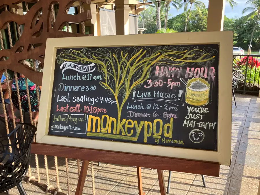 Chalkboard sign at Monkeypod Kitchen listing happy hour from 3:30 to 5PM, live music and dinner times