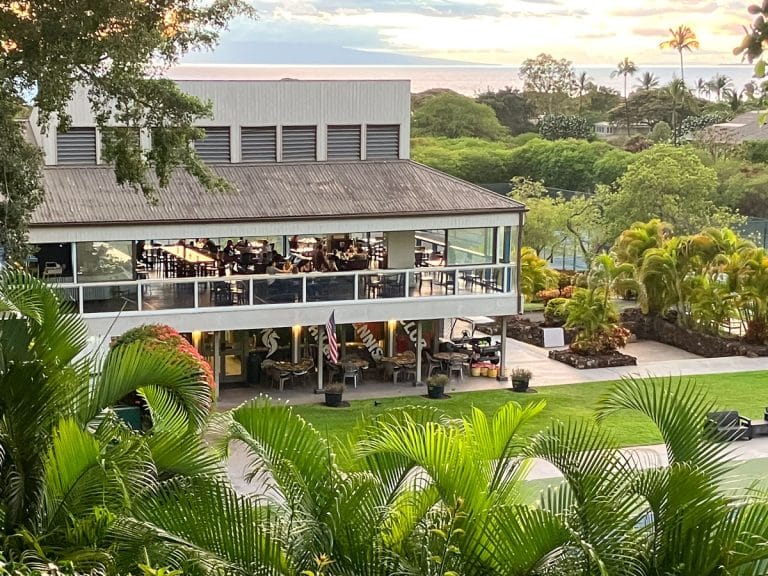 Diners on the upper-level lanai at Koast in Wailea, Maui, with palms in front and the ocean beyond at sunset
