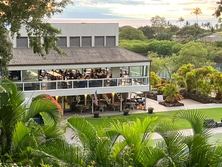 Diners on the upper-level lanai at Koast in Wailea, Maui, with palms in front and the ocean beyond at sunset
